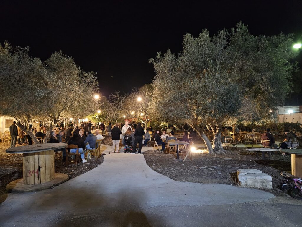 הכניסה לבר היין הקהילתי בקיבוץ רעים. בתמונה רואים את בר היין מלא באנשים באחד מערבי הקהילה בקיבוץ עם הופעות עמדות אוכל ושתייה שונות ומפגשי חברה